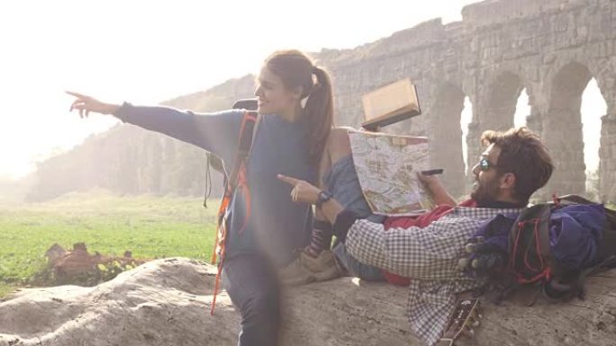 快乐的年轻夫妇背包客游客坐在原木后备箱上阅读地图指南指向罗马帕尔科德格里阿奎多蒂公园的古罗马渡槽遗址