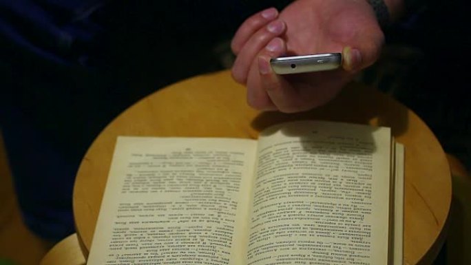 一名男子在纸质书的智能手机页面上拍照。打开的书放在椅子上。这个人翻过一页。