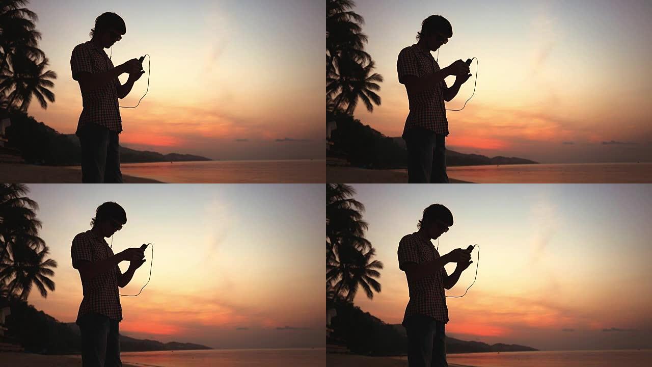 在美丽的日落时分，年轻的帅哥在热带海滩上戴着耳机和手机听音乐。慢动作。1920x1080