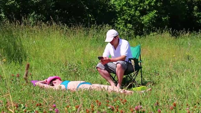 一个穿着泳衣的女孩正在草坪上晒日光浴。在她旁边的野餐椅上，一个男人坐在智能手机上玩耍。