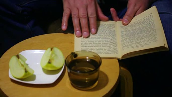 一个退休年龄的人正在读书。翻阅页面。他从杯子里喝水。附近是一个装有苹果片的碟子。