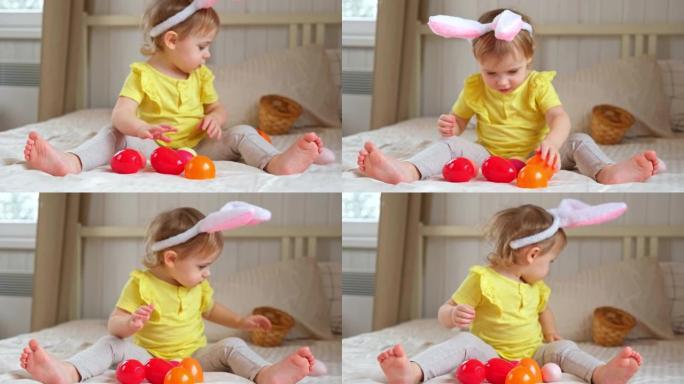 有兔子耳朵的女婴。可爱的搞笑宝宝在家玩复活节彩蛋。孩子在室内玩五颜六色的鸡蛋。春。用节日和宗教概念的