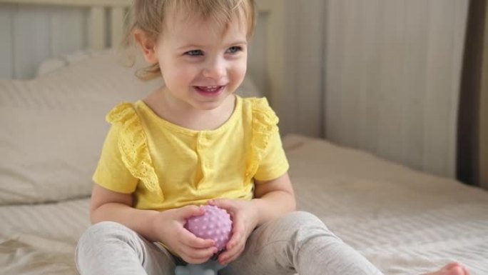 可爱的女婴坐在床上玩五颜六色的感官球。一岁的孩子玩触觉按摩球，以增强认知和物理过程。儿童发展，健康。