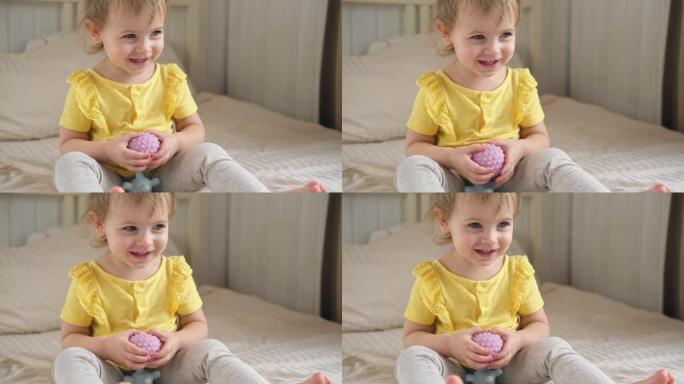 可爱的女婴坐在床上玩五颜六色的感官球。一岁的孩子玩触觉按摩球，以增强认知和物理过程。儿童发展，健康。