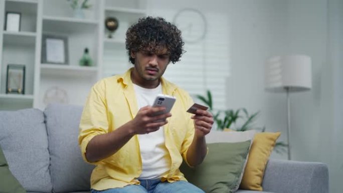 不成功的支付互联网诈骗受害者心烦意乱的印度人坐在家里的沙发上拿着卡和智能手机手机手掌感到震惊