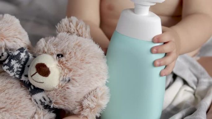 婴儿男孩孩子躺在床上，带着猫头鹰泰迪熊玩具和洗发水沐浴露瓶。蹒跚学步的婴儿肖像身体护理。学龄前儿童捡