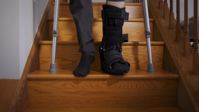 一个穿着黑色骨折靴的断腿男子试图用拐杖在客厅的楼梯上行走。