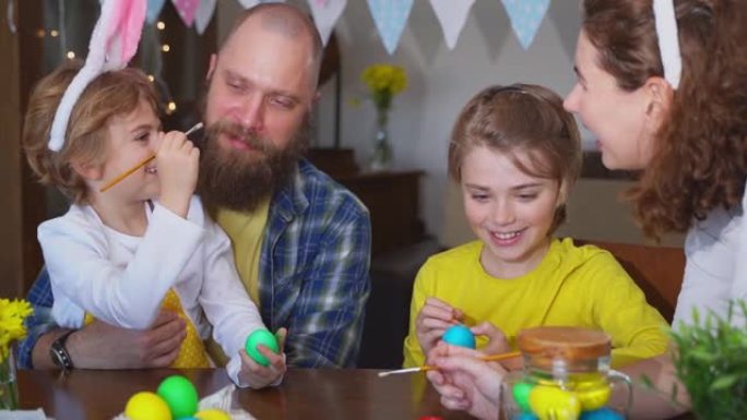 复活节家庭传统。父母和两个白种人快乐的孩子，兔子耳朵在一起坐在家里的桌子上，用油漆染和装饰鸡蛋。孩子