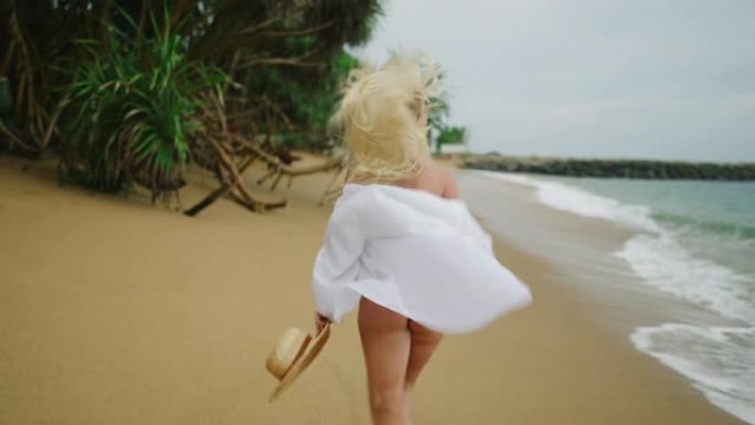 年轻的金发女子手持草帽，穿着白衬衫，沿着海滩逃离相机。白发的高加索女孩随风在海上奔跑，转向相机
