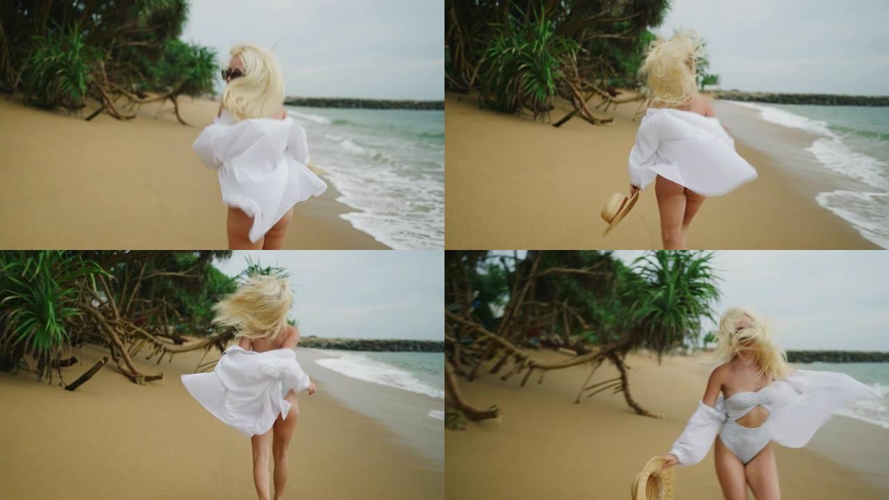 年轻的金发女子手持草帽，穿着白衬衫，沿着海滩逃离相机。白发的高加索女孩随风在海上奔跑，转向相机