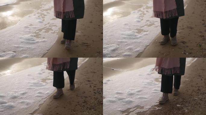女人穿着靴子沿着风沙在沙滩上吹来的海泡沫以慢动作行走。在风雨如磐的日子里，穿着防水鞋的女性脚在海浪的