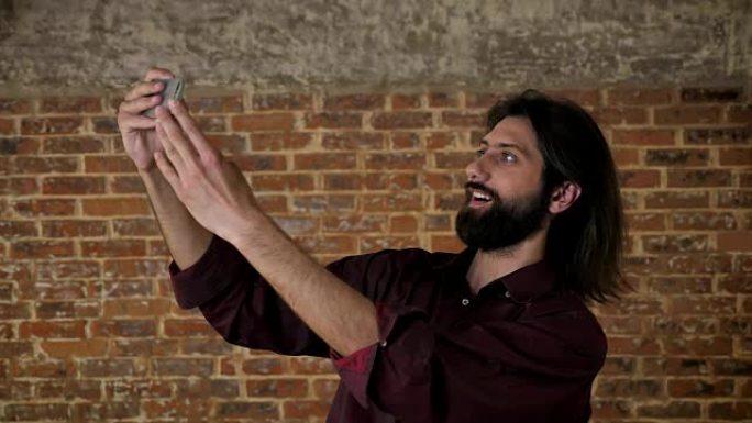 年轻迷人的黑发胡子男人正在智能手机上制作视频，鬼混，砖块背景，交流概念