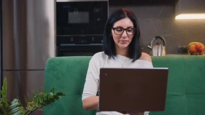 坐在绿色沙发上的迷人的沉思黑发女人的特写镜头在笔记本电脑上远程工作，在家做自由职业在线工作。工作流概