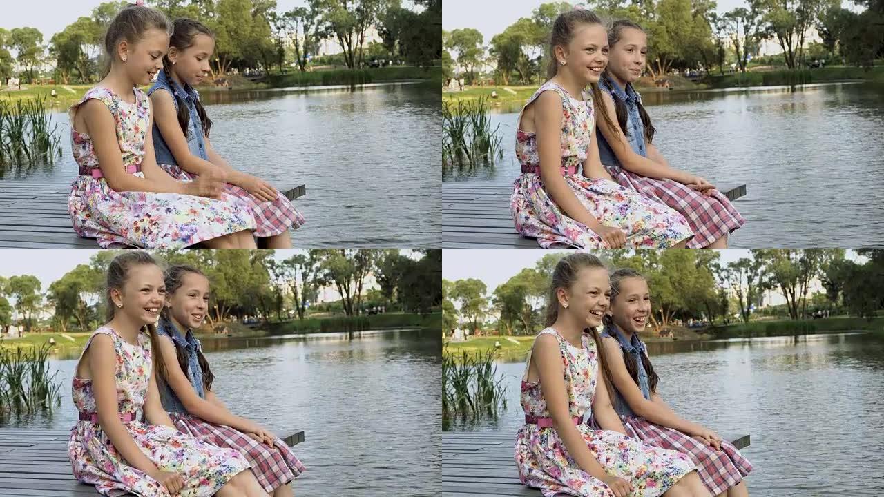 两个开朗的小女孩在城市公园的河边坐在夏天的木桥上，彼此相邻，看着一边笑。肖像。