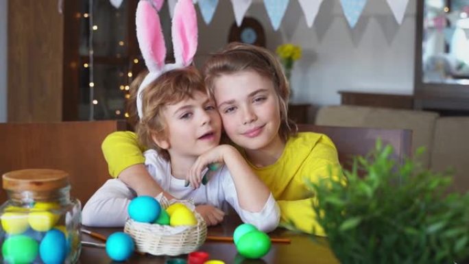 复活节周日家庭节日传统。两个白种人快乐的兄弟姐妹孩子，兔子耳朵摆姿势微笑，看着相机装饰的鸡蛋，坐在家