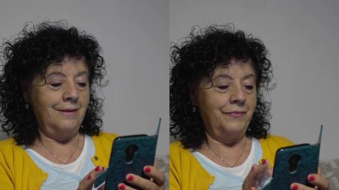 一个穿着黄色毛衣的老妇人坐在客厅沙发上，一边看着并操纵着她的带书皮的手机的垂直视频。