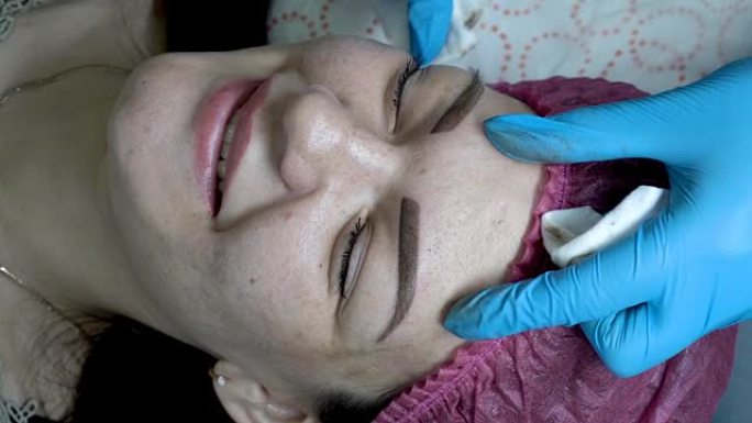 美容师在永久化妆眉毛的程序后，在美容院中治疗女孩眉毛周围的皮肤。非常特写。从顶部看。4K，25 fp