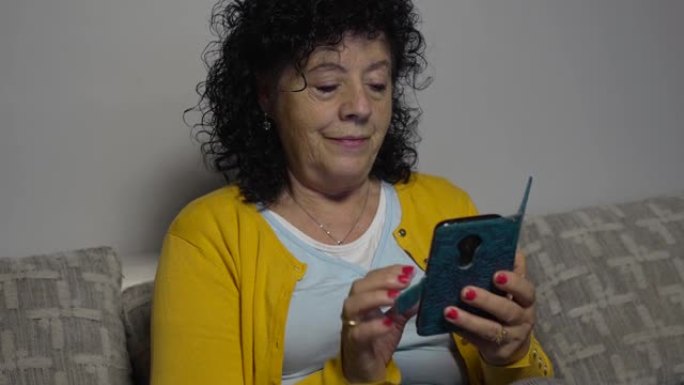 一个穿着黄色毛衣的老妇人坐在客厅沙发上，看着并操纵着她的带书皮的手机的视频。