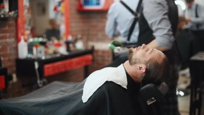 工人在顾客脸上撒水并清洗。长发的时尚男人在理发店开会