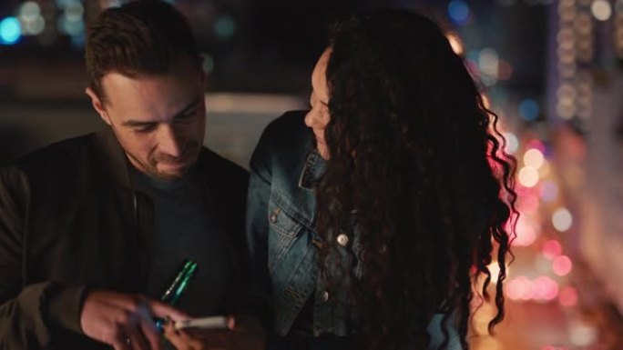 快乐的高加索夫妇晚上在屋顶上放松，使用智能手机浏览照片，晚上在城市享受浪漫的约会