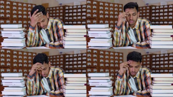 穿着便服的疲惫的印度学生坐在书屋的书桌前，双手放在头上，看起来厌倦了阅读