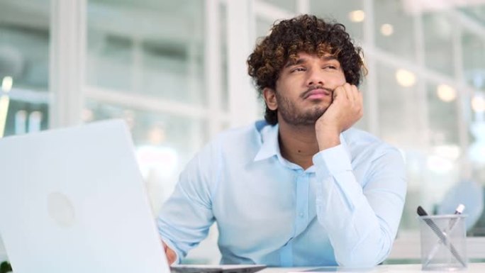 劳累过度的年轻工人无聊地坐在现代办公室的办公桌前。穿着衬衫的精疲力尽的男人因使用笔记本电脑而感到疲倦