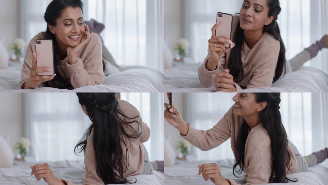 美丽的女人用智能手机视频聊天，和朋友聊天，享受炫耀躺在床上放松在家的公寓