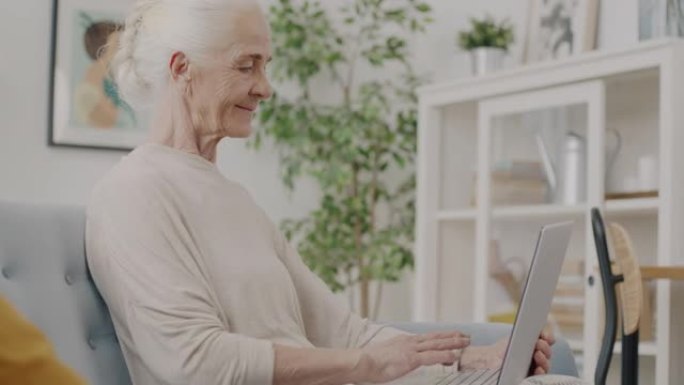 退休女士的慢动作与笔记本电脑一起工作，在家中使用无线技术打字微笑