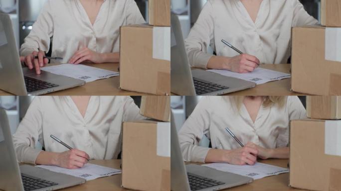 一名年轻女子写退货单，坐在家里有笔记本电脑的桌子旁。一位女顾客在纸上写字并填写表格，想要退回纸板箱的