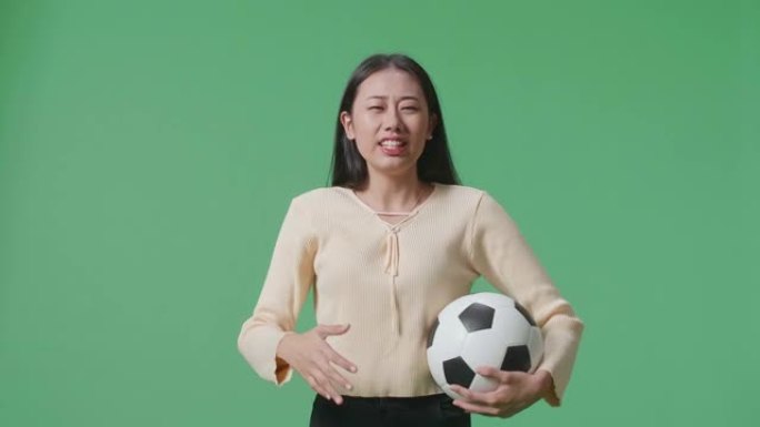 亚洲女子带球正在为射门而生根，并在绿屏背景上欢呼足球时后悔
