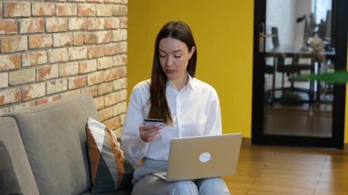 慢动作视频白衬衫和灰色裤子的白人美女正坐在灰色沙发上，在电脑上工作。