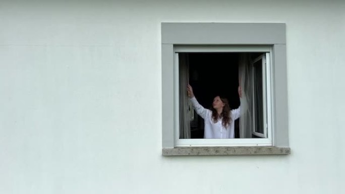 一个十几岁的女孩劈开白色的窗帘，看着白色的窗户。她在视频的右侧。在左侧，有很多文字空间。她向外看并微