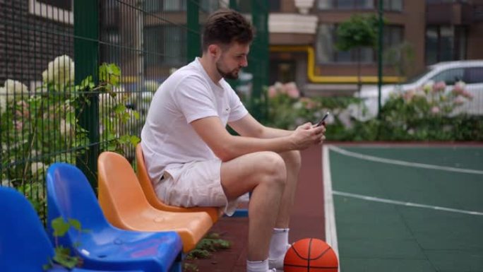 自信的运动员在城市的运动场上拥有智能手机和户外球。微笑满意的白人男子在网上传递训练中断的肖像。慢动作
