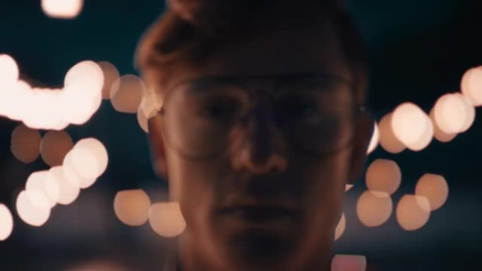 肖像年轻的红发男子戴眼镜在屋顶上晚上看着严肃的表情与美丽的派对灯背景的城市青年概念