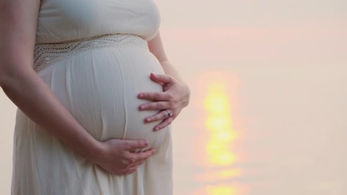 一名孕妇站在床后的海边，特写镜头，抚摸着她的肚子