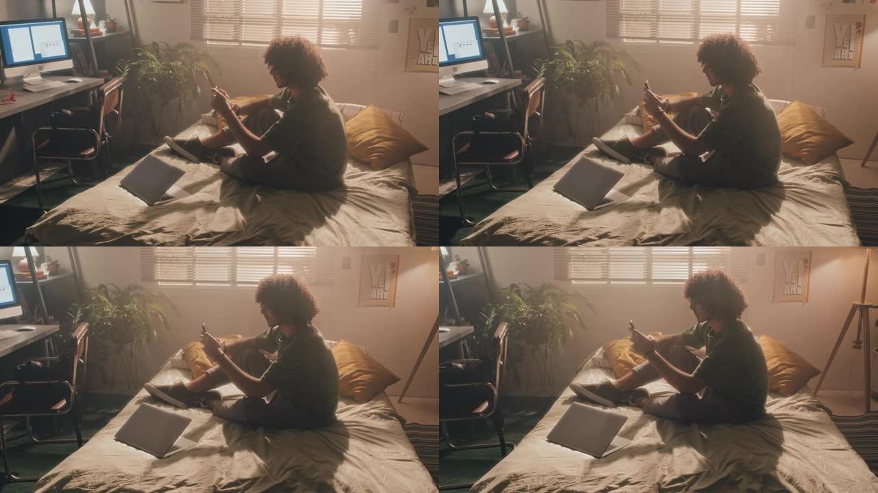 卷曲的年轻人坐在未整理的床上，阳光充足的卧室里，在手机上打字