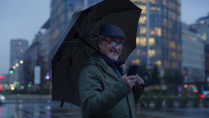 中年男子站在雨下的街道上，拿着雨伞，使用智能手机的肖像。高级男子发短信，聊天，在旅途中使用技术进行地