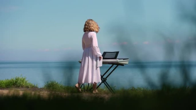 河岸上的钢琴合成器，十几岁的高加索女孩进入慢动作走向乐器。自信的有创造力的音乐家坐在风景的背景下。