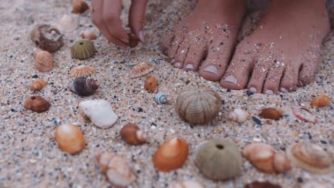 特写脚女人在海滩上收集贝壳，享受美丽的自然品种，在沙滩上塑造图案
