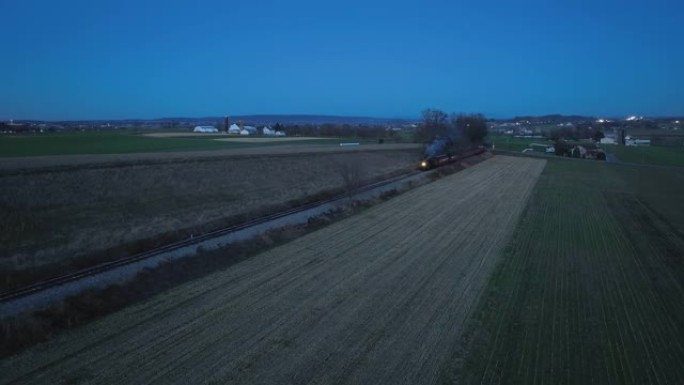 一辆蒸汽旅客列车在一条穿过农田的单轨轨道上驶近的无人机夜景