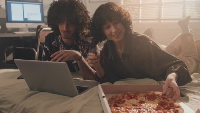 年轻的混血儿夫妇躺在床上在笔记本电脑上看电影，吃披萨