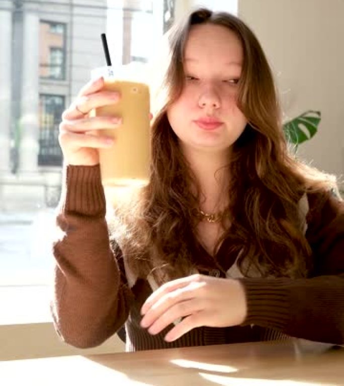 一个十几岁女孩的特写嘴唇在玻璃杯里喝卡布奇诺冷咖啡和牛奶棕色夹克阁楼风格的脖子上的链子装饰美味的咖啡