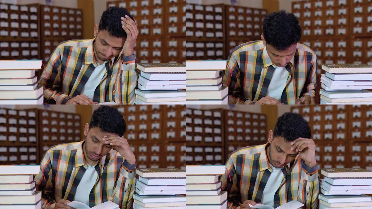 穿着便服的疲惫的印度学生坐在书屋的书桌前，双手放在头上，看起来厌倦了阅读