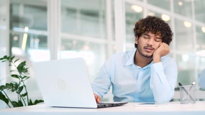 劳累过度的年轻工人感到疲倦，想坐在现代办公室的办公桌前工作时睡在笔记本电脑上。困得精疲力竭的男人穿着