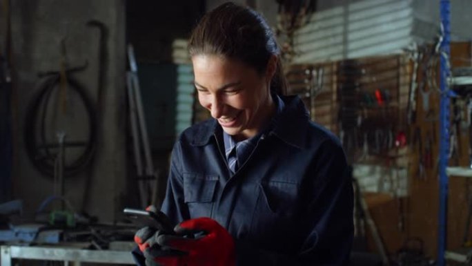 穿着制服工作服的年轻女机械师的肖像，使用智能手机与家人和朋友聊天。从事焊工工作的职业妇女在维修车间休