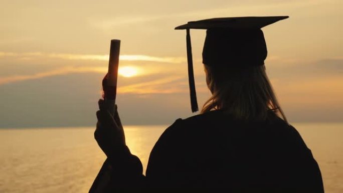 一个男人手里拿着文凭，一个毕业生的衣服看着海上的日出