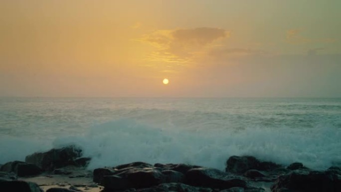 海洋上空地平线上昏黄的太阳。日落橙色的天空，光线在海水上反射。巨浪在岩石上破裂，飞溅飞入相机。