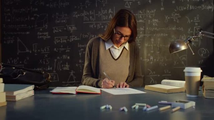 大学教室里年轻女教师的肖像，用数学公式在黑板前的桌子上工作。在大学图书馆学习的理科生看着相机微笑