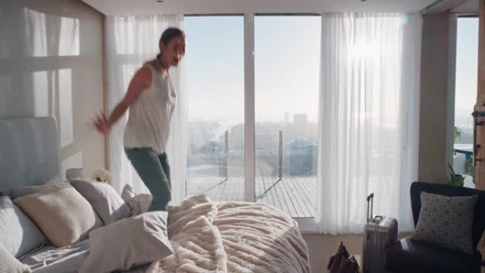 快乐的女人在酒店房间的床上蹦蹦跳跳，享受成功的生活方式，庆祝享受豪华的顶层公寓