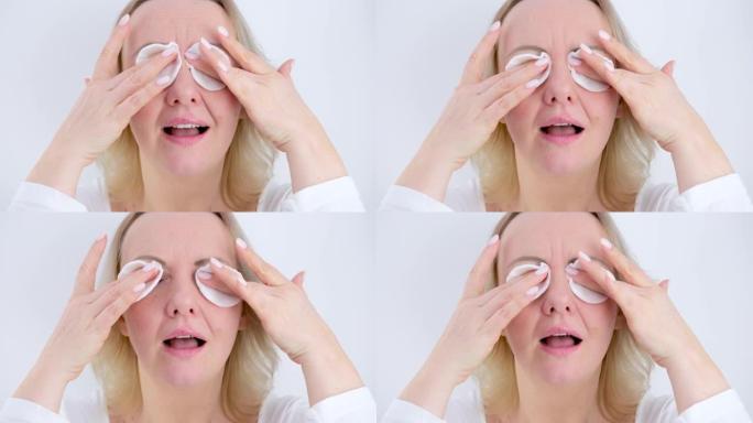 一名妇女将海绵放在眼睛上并摩擦手指卸妆洗掉化妆品晚上的日常程序在白色背景上化妆后的白色修指甲中年妇女